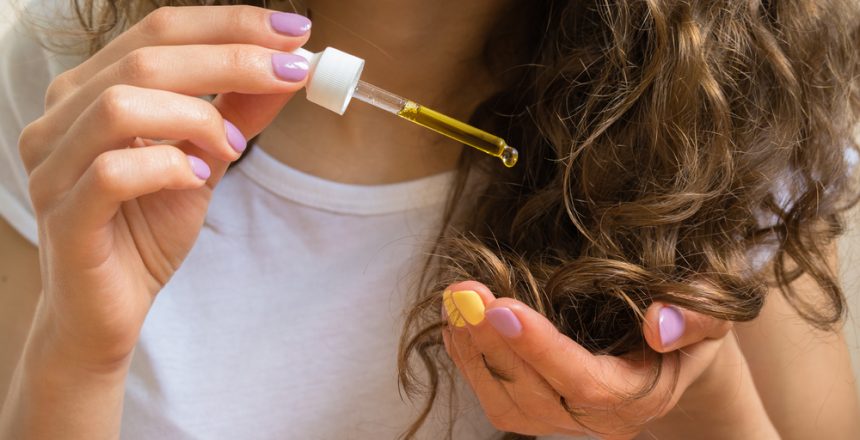 Young girl in white t-shirt holds in her left hand the tips of her curly hair and her right hand applies them yellow oil.